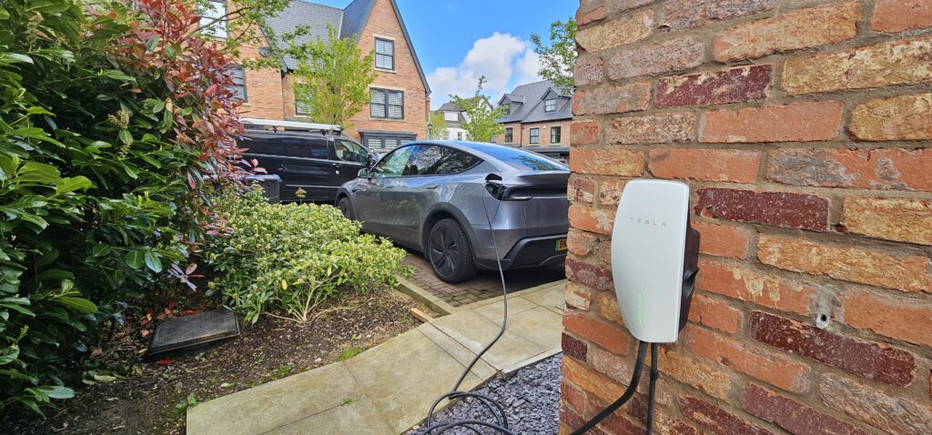 20260418_103535 Grey Tesla with a charging cable plugged into a wall charger mounted on a brick exterior in a driveway with a black van and greenery nearby